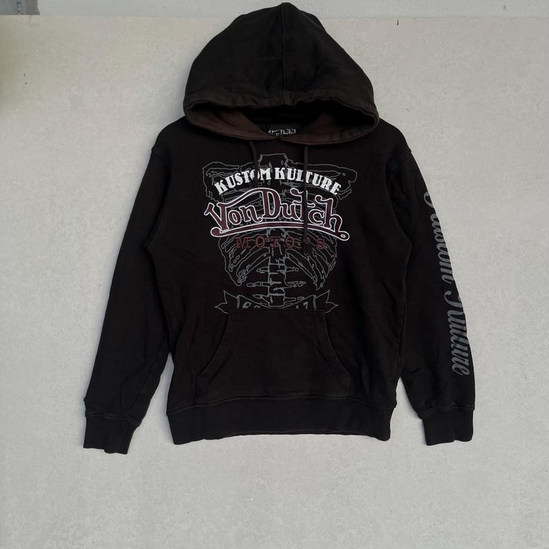 May include: A black long-sleeve hooded sweatshirt featuring a brown inner hood lining and a front graphic print. The design includes the text KUSTOM KULTURE and Von Dutch, along with a skeletal rib cage illustration and the word MOTORS in red.