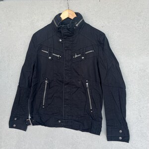 Puede incluir: Chaqueta negra con m&uacute;ltiples cremalleras plateadas y detalles de botones. La chaqueta tiene un cuello alto y una silueta entallada. Cuelga de una percha de madera. La chaqueta parece estar hecha de una tela duradera.