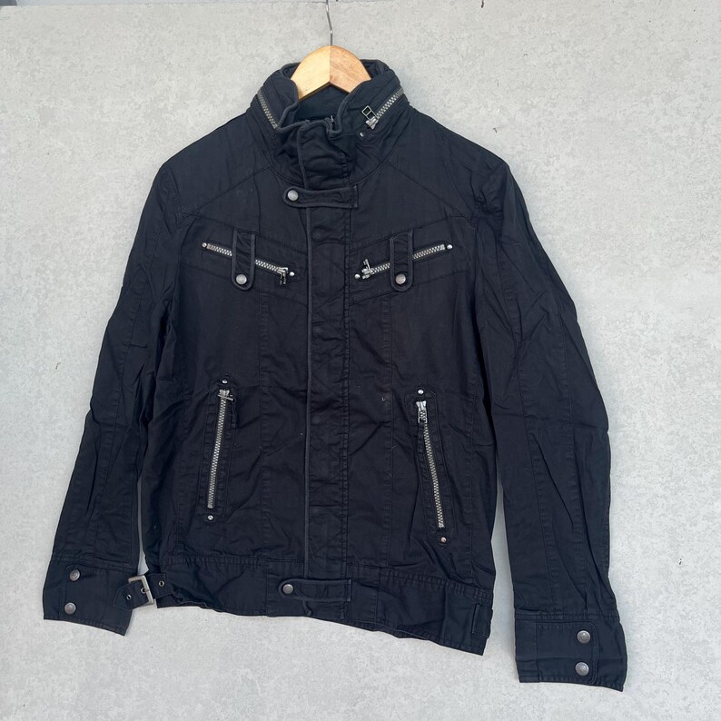 Puede incluir: Chaqueta negra con m&uacute;ltiples cremalleras plateadas y detalles de botones. La chaqueta tiene un cuello alto y una silueta entallada. Cuelga de una percha de madera. La chaqueta parece estar hecha de una tela duradera.
