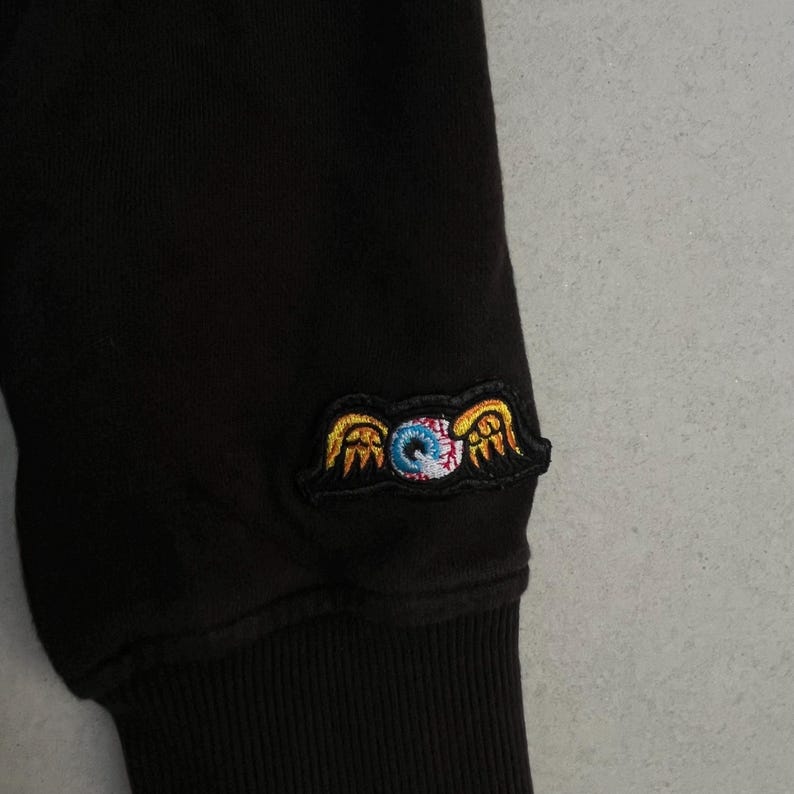 May include: A black long-sleeved garment featuring an embroidered patch on the sleeve. The patch depicts a central blue and white eyeball with red veins, flanked by two stylised yellow wings with orange outlines, all set against a black background.