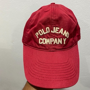 May include: A red baseball cap with the text "POLO JEANS COMPANY" in white lettering. The cap has a curved brim and a fabric strap on the back for adjustment. The hat is made of a soft, durable material.