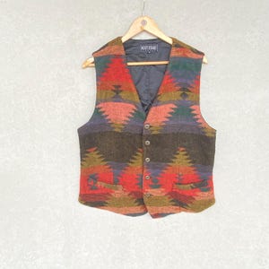 May include: A Next Stage vest with a vibrant geometric pattern in red, orange, green, and purple. The vest features a button-up front, two pockets, and a black lining. It is displayed on a wooden hanger.