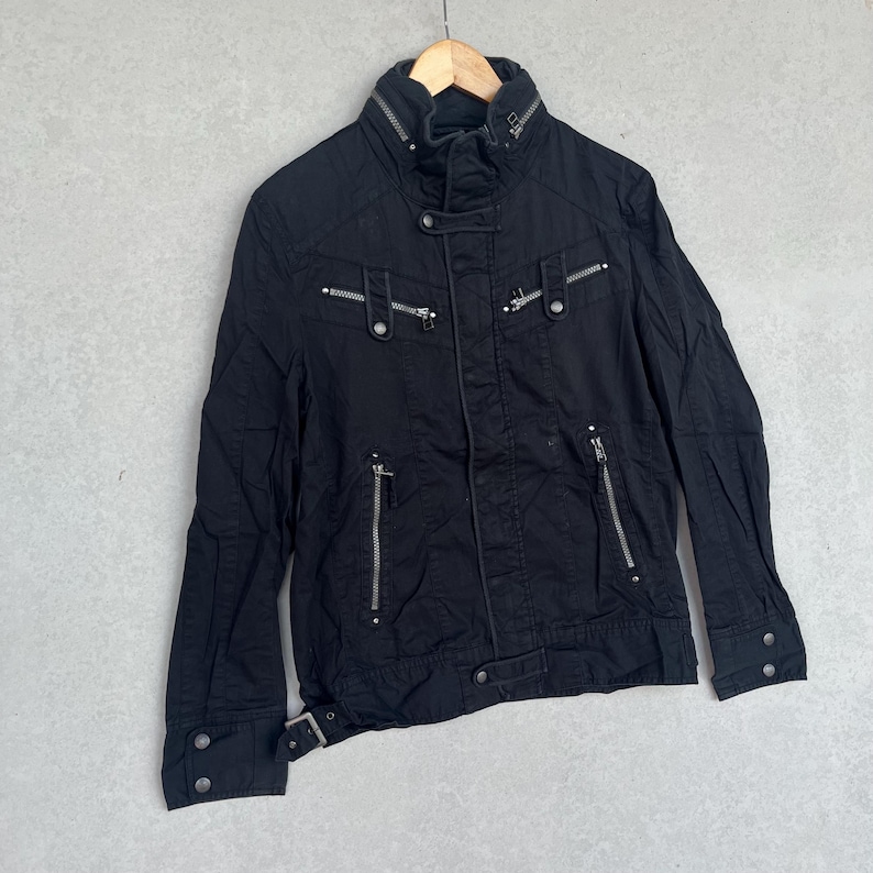 Puede incluir: Chaqueta negra con cuello alto, m&uacute;ltiples bolsillos con cremallera y detalles plateados. La chaqueta presenta un detalle de cintur&oacute;n en la cintura y pu&ntilde;os abotonados. Cuelga de una percha de madera sobre un fondo gris neutro.