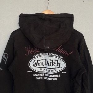 May include: A black hooded sweatshirt featuring a large white oval graphic on the back with the text Von Dutch Motors, Pinstriping Specialist, Master Mechanics, and West Coast.US. The design includes distressed lettering and a hood.