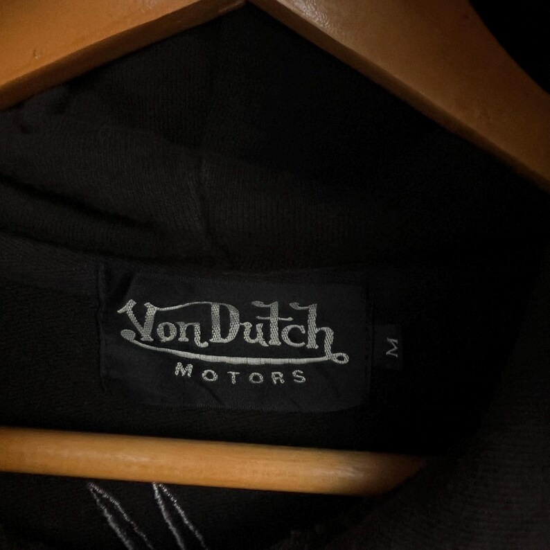 May include: A black hooded sweatshirt featuring a rectangular fabric label sewn into the inner neckline. The label displays the white embroidered text Von Dutch MOTORS with a stylised underline and a separate small tag indicating size M.