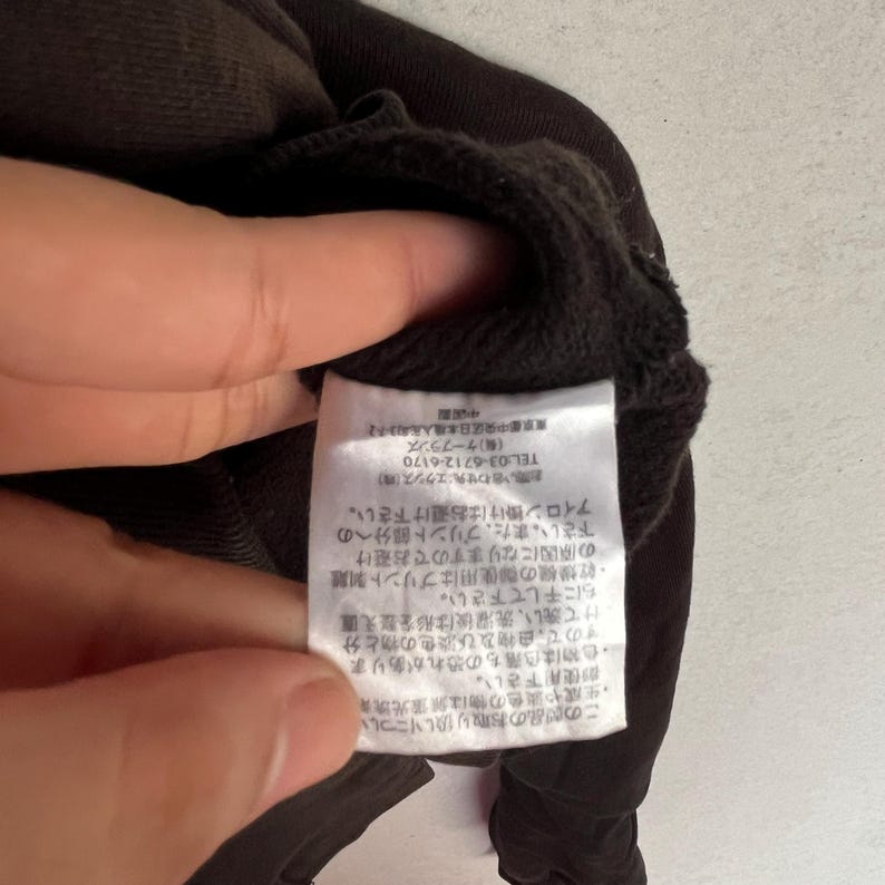 May include: A dark brown garment featuring a white rectangular care label sewn into the interior seam. The label contains detailed Japanese text instructions regarding washing, drying, and maintenance, along with contact information for the manufacturer.
