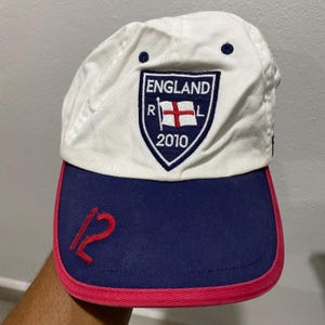May include: White baseball cap with a navy blue brim and red trim. The cap features an embroidered shield with the text "ENGLAND R L 2010" and a cross flag. A red number "12" is embroidered on the side.
