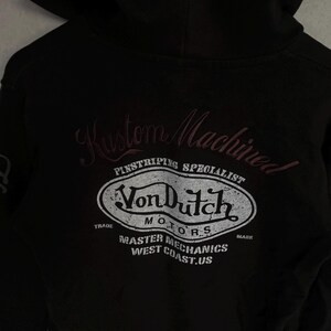 May include: A black hooded sweatshirt featuring a distressed white graphic on the back. The text reads Kustom Machined, Pinstriping Specialist, Von Dutch Motors, Trade Mark, Master Mechanics, and West Coast.US in a stylised oval emblem design.