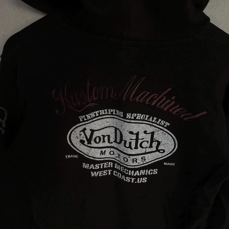 May include: A black hooded sweatshirt featuring a distressed white graphic on the back. The text reads Kustom Machined, Pinstriping Specialist, Von Dutch Motors, Trade Mark, Master Mechanics, and West Coast.US in a stylised oval emblem design.