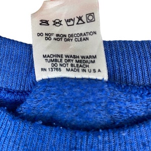 May include: A white care tag with blue text detailing washing instructions for a garment. The tag reads "40% DO NOT IRON DECORATION DO NOT DRY CLEAN MACHINE WASH WARM TUMBLE DRY MEDIUM DO NOT BLEACH RN 13765 MADE IN USA".