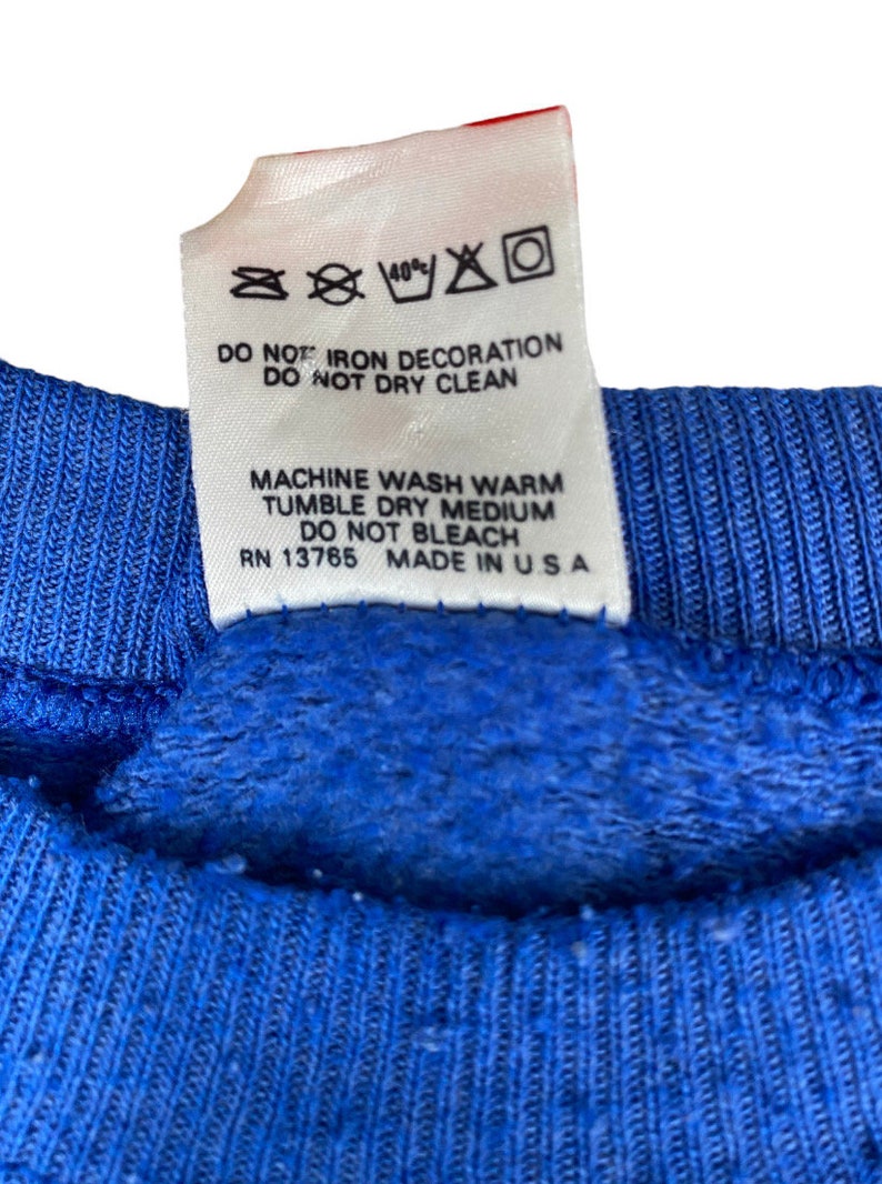 May include: A white care tag with blue text detailing washing instructions for a garment. The tag reads "40% DO NOT IRON DECORATION DO NOT DRY CLEAN MACHINE WASH WARM TUMBLE DRY MEDIUM DO NOT BLEACH RN 13765 MADE IN USA".