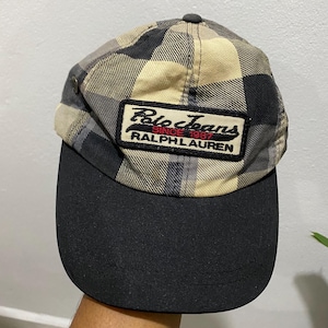 May include: A plaid baseball cap with a black brim. The cap features a patch with the text "Polo Jeans Since 1967 Ralph Lauren" in red and white. The cap is a mix of black, tan, and gray.