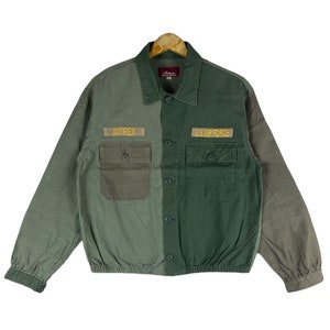 May include: A green and brown cotton jacket with a button-up front. The jacket has two pockets on the front and features the words "AVIREX" and "U.S. AIR FORCE" embroidered on the pockets.