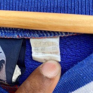 May include: Close-up of a blue garment with a white tag. The tag has Japanese text and numbers. The garment is blue with white and red accents. A wooden hanger is visible at the top of the image.