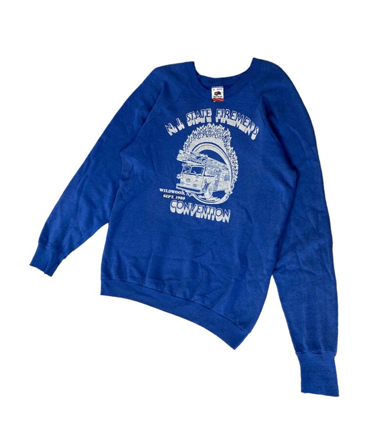 May include: A blue sweatshirt with a white graphic of a fire truck and the text "N.J. State Firemen's Convention" and "Wildwood Sept. 1989".