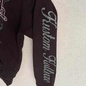 May include: A black long-sleeved hooded sweatshirt featuring the text Kustom Kulture printed in a grey, stylised script font vertically down the length of the sleeve. The garment includes a partial graphic design on the chest area.