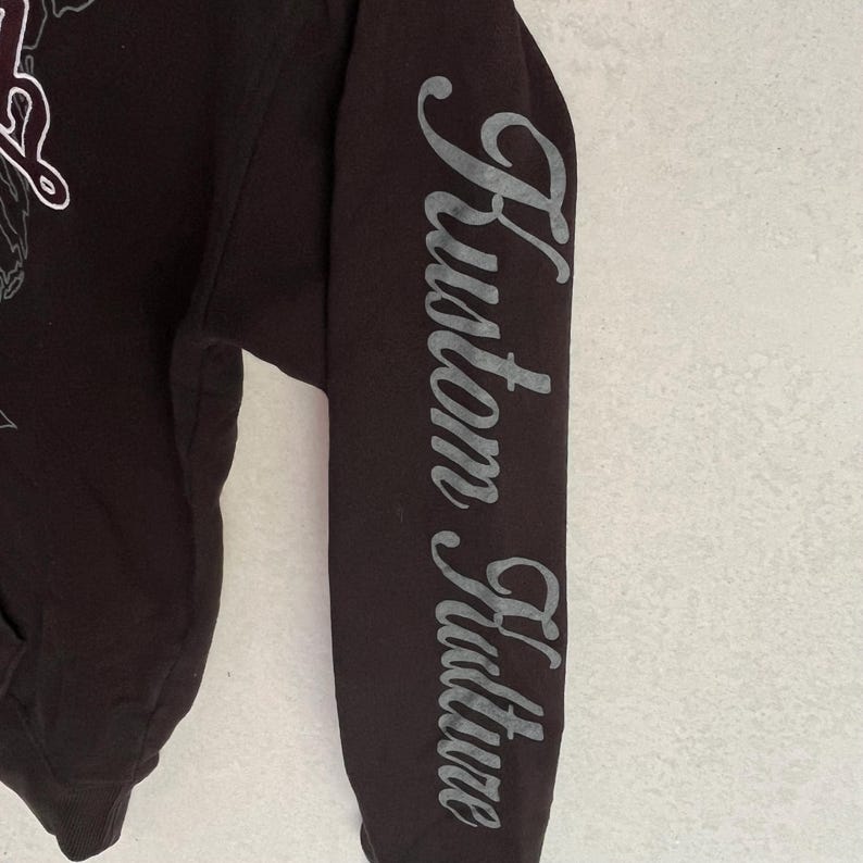 May include: A black long-sleeved hooded sweatshirt featuring the text Kustom Kulture printed in a grey, stylised script font vertically down the length of the sleeve. The garment includes a partial graphic design on the chest area.