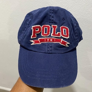 May include: Navy blue baseball cap with the word "POLO" in white and red lettering. The year "1967" is embroidered below the word "POLO" on a red banner. The cap has a curved brim and is made of a soft fabric.