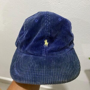May include: A navy blue corduroy baseball cap with a curved brim. The cap features a small, embroidered yellow logo of a polo player on the front. The hat has a classic six-panel design and a slightly worn appearance.