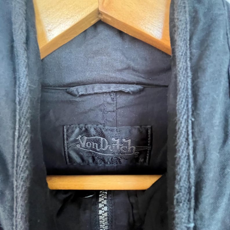 Puede incluir: Primer plano de una chaqueta negra con cremallera y capucha. El logotipo de Von Dutch es visible en una etiqueta negra. La chaqueta est&aacute; colgada en una percha de madera.