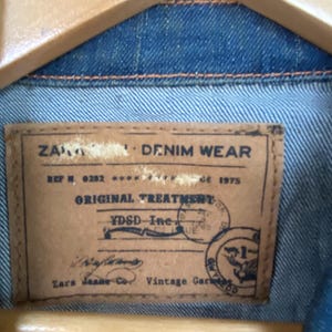 Pu&ograve; includere: Primo piano dell'interno di una giacca di jeans, che mostra un'etichetta in pelle marrone. L'etichetta recita "ZARA DENIM WEAR" con dettagli come "ORIGINAL TREATMENT" e "Vintage Garment". La giacca &egrave; blu.