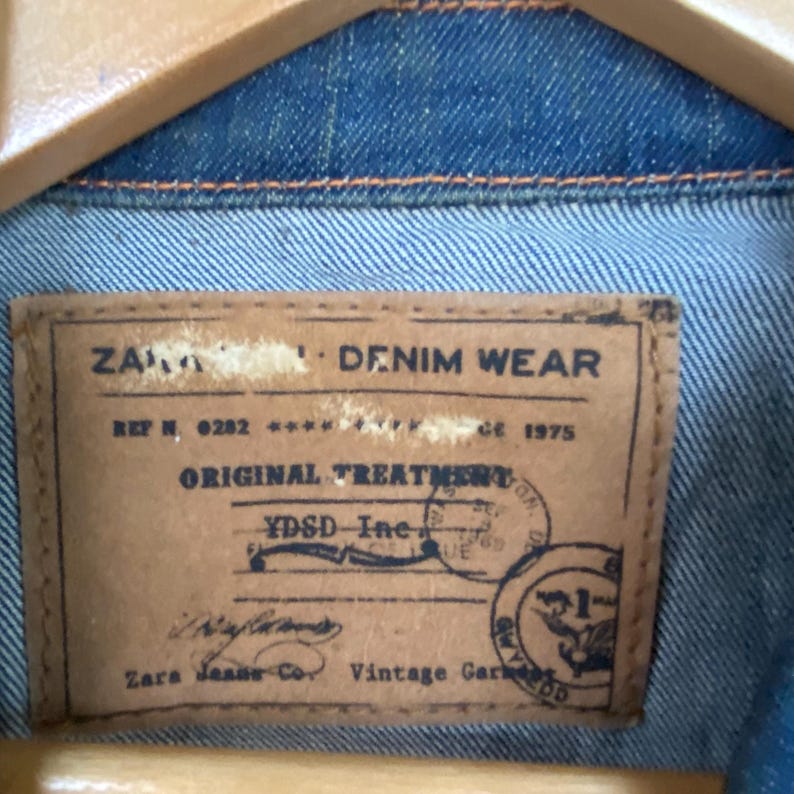 Pu&ograve; includere: Primo piano dell'interno di una giacca di jeans, che mostra un'etichetta in pelle marrone. L'etichetta recita "ZARA DENIM WEAR" con dettagli come "ORIGINAL TREATMENT" e "Vintage Garment". La giacca &egrave; blu.