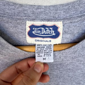 May include: A heather gray t-shirt featuring a white fabric tag with a blue shield-shaped logo that reads Von Dutch ORIGINALS. Below the logo, a secondary tag lists care instructions, 100% cotton, Made in U.S.A., and the size M.