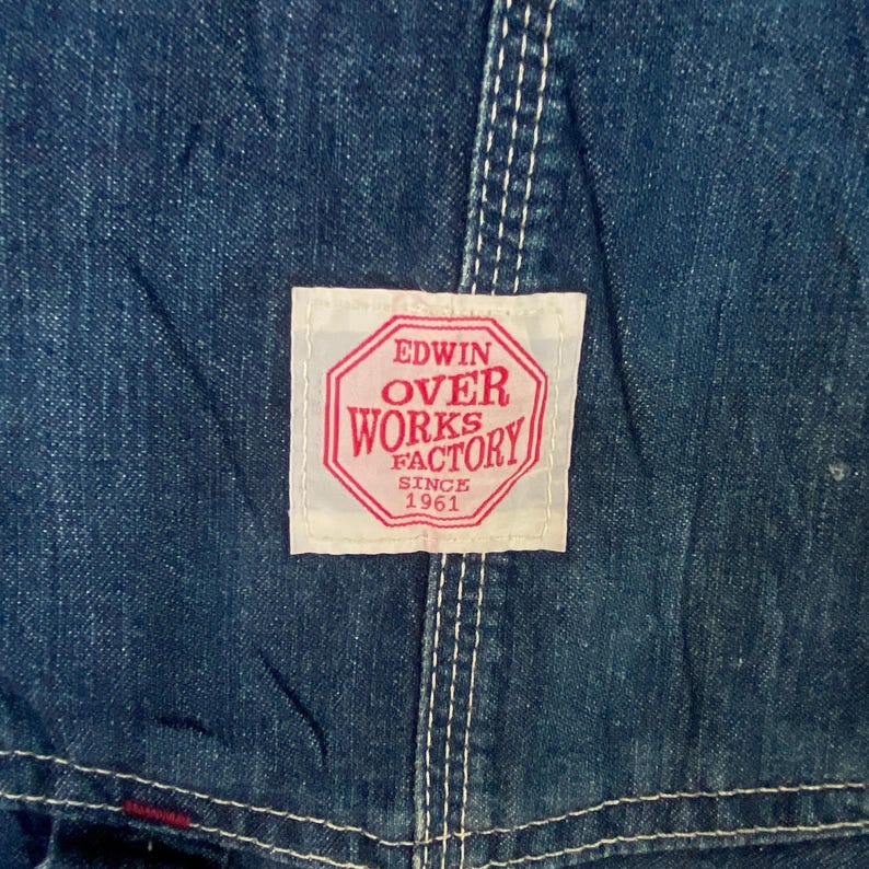 May include: Close-up of blue denim fabric with white stitching. A rectangular patch with red text reads "EDWIN OVER WORKS FACTORY SINCE 1961". The denim has a worn appearance.