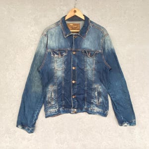 Pu&ograve; includere: Giacca di jeans blu dal design classico, con chiusura a bottoni, tasche sul petto e tasche laterali. La giacca &egrave; appesa a una gruccia di legno su uno sfondo neutro. Il denim ha un aspetto sbiadito e usurato.