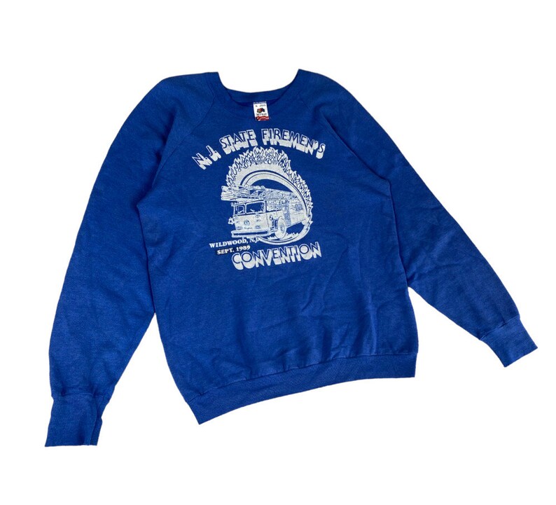 May include: A blue sweatshirt with a white graphic of a fire truck and the text "N.J. State Firemen's Convention" with the date "Sept. 1989" and location "Wildwood, N.J."