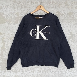 Puede incluir: Sudadera negra de Calvin Klein Jeans con un gran logotipo "CK" blanco en la parte delantera. El nombre de la marca está impreso en blanco debajo del logotipo. La sudadera está colgada en una percha de madera.