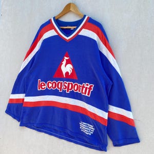 May include: Blue long-sleeve shirt with red and white accents. Features the text "le coq sportif" in white, along with a rooster logo. The shirt has a V-neck design and is hanging on a wooden hanger.
