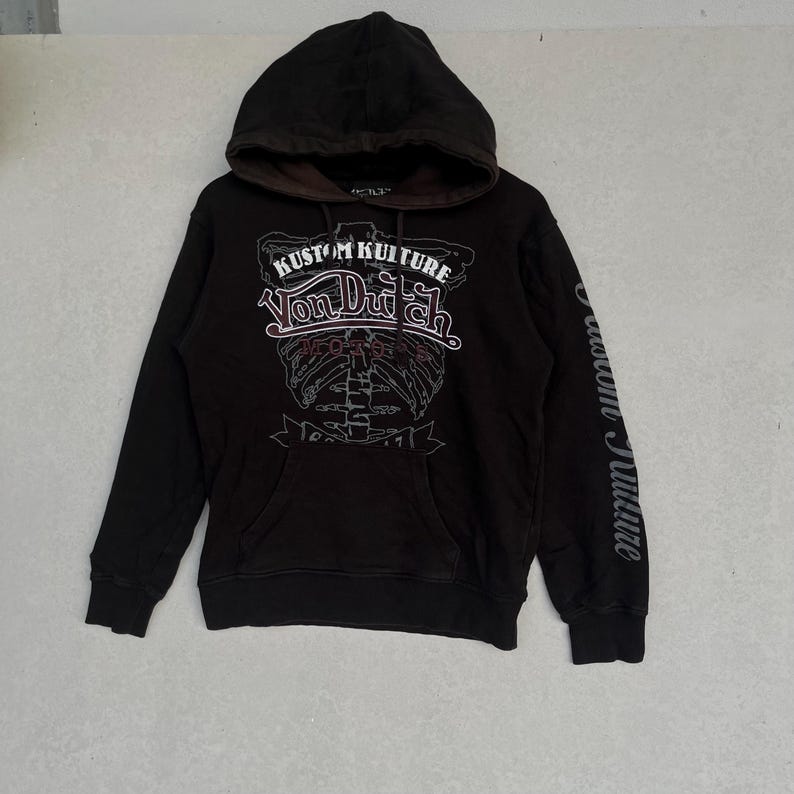 May include: A black hooded sweatshirt featuring a brown inner hood lining and a front graphic print that reads KUSTOM KULTURE VON DUTCH MOTORS. The design includes a skeletal rib cage illustration and the text KUSTOM KULTURE on the left sleeve.