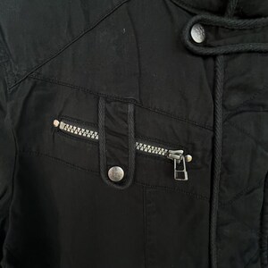 Puede incluir: Primer plano de una chaqueta negra con cremallera y un peque&ntilde;o bolsillo. La chaqueta tiene un dise&ntilde;o acolchado y una capucha con cord&oacute;n. La cremallera es plateada y hay un bot&oacute;n plateado en el bolsillo.