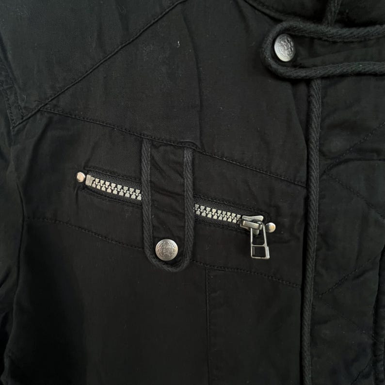 Puede incluir: Primer plano de una chaqueta negra con cremallera y un peque&ntilde;o bolsillo. La chaqueta tiene un dise&ntilde;o acolchado y una capucha con cord&oacute;n. La cremallera es plateada y hay un bot&oacute;n plateado en el bolsillo.