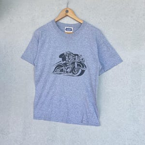 May include: A heather gray short-sleeve t-shirt featuring a dark navy blue graphic print of a person riding a motorcycle. The motorcycle has the text "VON DUTCH" printed on its side. The shirt includes a small brand label at the inner neckline.