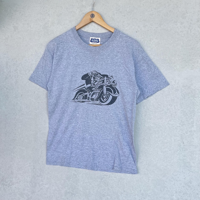 May include: A heather gray short-sleeve t-shirt featuring a dark navy blue graphic print of a person riding a motorcycle. The motorcycle has the text "VON DUTCH" printed on its side. The shirt includes a small brand label at the inner neckline.