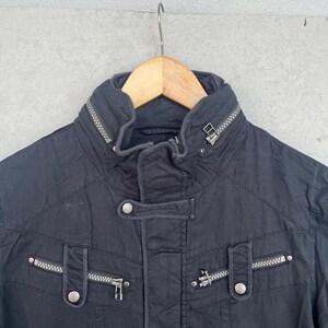 Puede incluir: Chaqueta negra con cuello alto y m&uacute;ltiples cremalleras. La chaqueta tiene cierre de bot&oacute;n y dos bolsillos con cremalleras. La chaqueta est&aacute; colgada en una percha de madera.