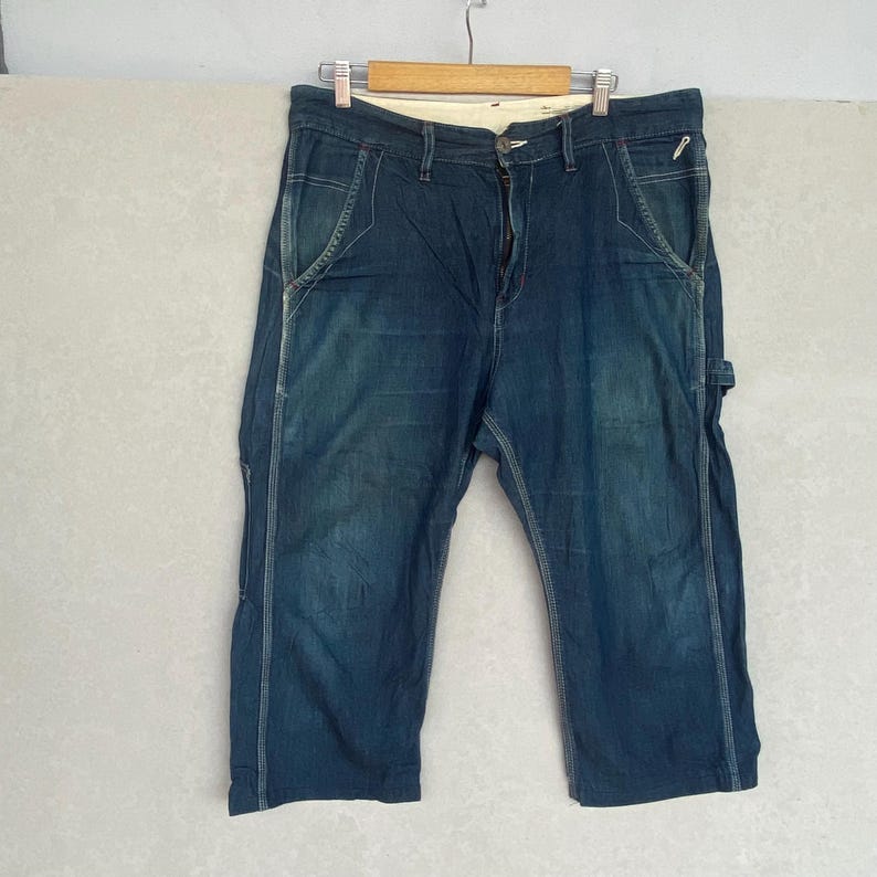 May include: Dark wash denim carpenter shorts with white stitching and a button and zip closure. The shorts have a relaxed fit and feature multiple pockets and a hammer loop. They are hanging on a wooden hanger.