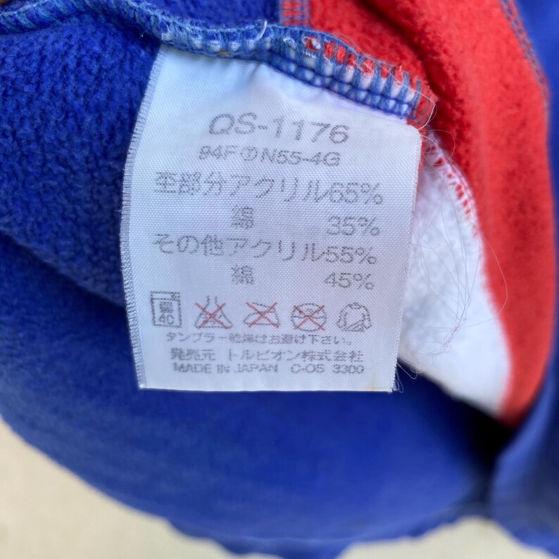 May include: Close-up of a white tag with Japanese text and care symbols. The tag is attached to a blue, red, and white garment. Text includes "QS-1176", "94FN55-4G", and fabric composition percentages. The tag also has washing instructions.