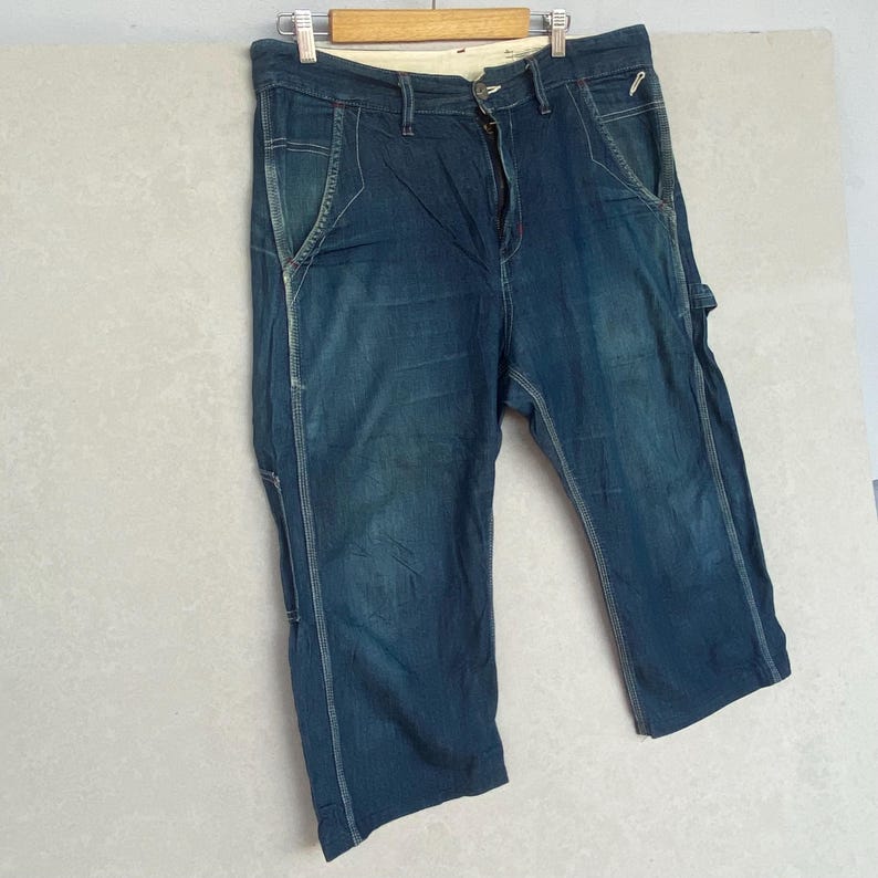 May include: Blue denim carpenter trousers with white stitching and multiple pockets. The trousers are hanging on a wooden hanger. The denim has a faded appearance, suggesting wear. The trousers have a button and zip closure.
