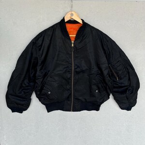 May include: Black bomber jacket with a zip-up front, two front pockets, and a ribbed collar and cuffs. The jacket has an orange lining and is hanging on a wooden hanger. A classic outerwear piece.