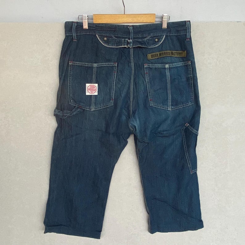 May include: Dark blue denim capri trousers with multiple pockets and a belt loop detail. The back features two pockets with red stitching and a patch with text. The text "OVER WORKS FACTORY" is visible on a label.