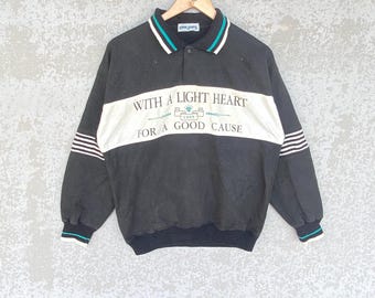 May include: Black and white long-sleeved sweatshirt with a collared neckline. The shirt features the text "WITH A LIGHT HEART FOR A GOOD CAUSE" across the chest. The collar and cuffs have teal and white accents.