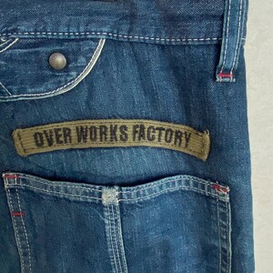 May include: Close-up of blue denim jeans with a back pocket and a brown patch that reads "OVER WORKS FACTORY". The jeans have white stitching and a metal button. The denim has a faded appearance.