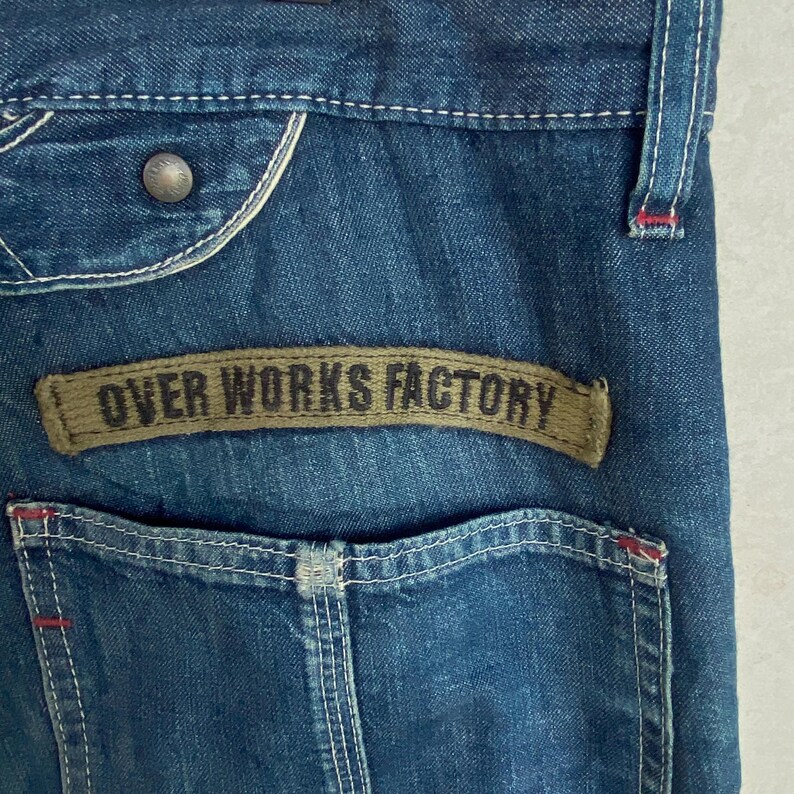 May include: Close-up of blue denim jeans with a back pocket and a brown patch that reads "OVER WORKS FACTORY". The jeans have white stitching and a metal button. The denim has a faded appearance.