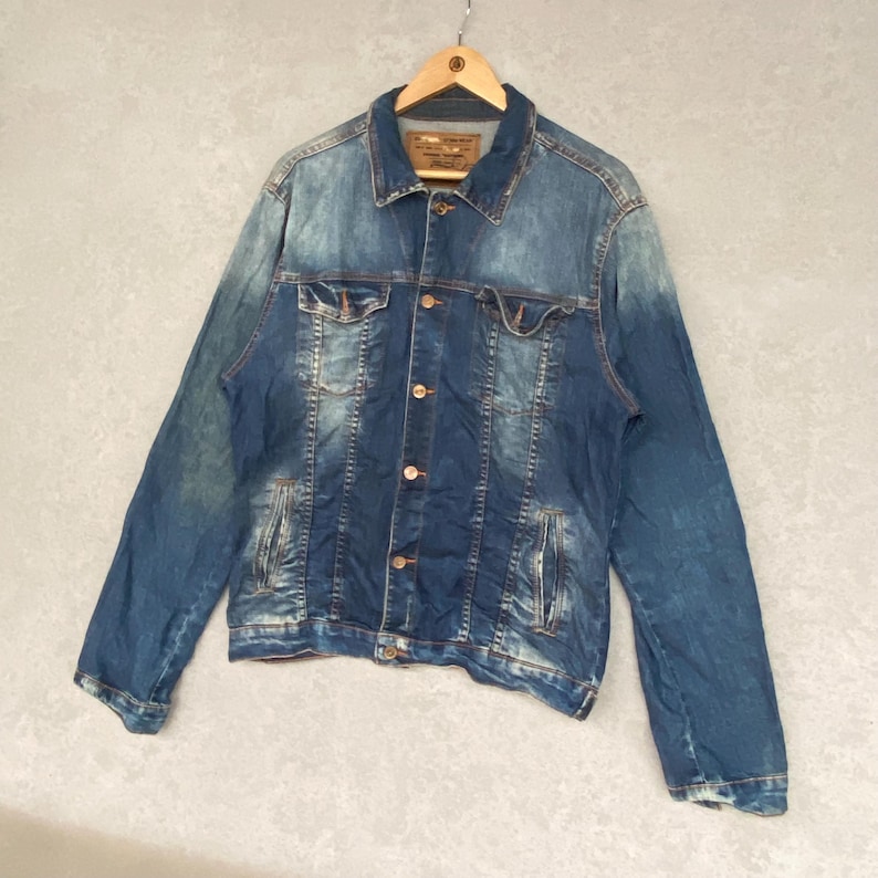 Pu&ograve; includere: Giacca di jeans blu dal design classico, con chiusura a bottoni, tasche sul petto e tasche laterali. La giacca &egrave; appesa a una gruccia di legno su uno sfondo neutro. Il denim ha un aspetto sbiadito e usurato.