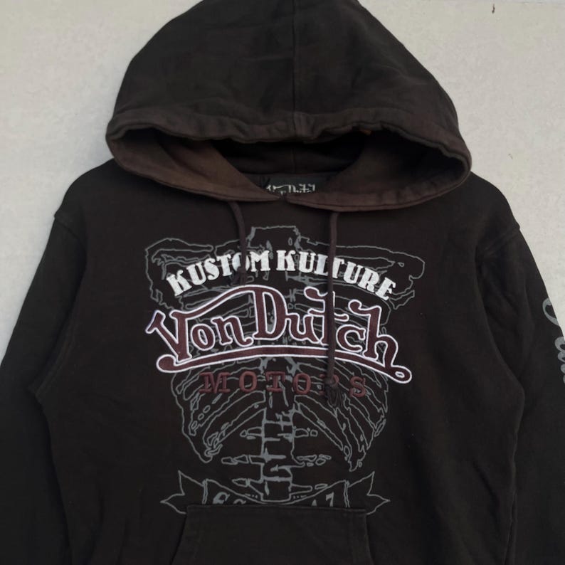 May include: A dark brown hooded sweatshirt featuring a large graphic print on the chest. The design includes a white skeleton rib cage outline, the text KUSTOM KULTURE, the Von Dutch logo in white and maroon, and the word MOTORS in maroon lettering.