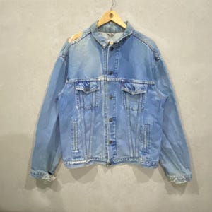 May include: Light blue denim jacket with button closures, two chest pockets, and two side pockets. The jacket is hanging on a wooden hanger. The denim has a faded appearance, suggesting it is vintage or has been washed.