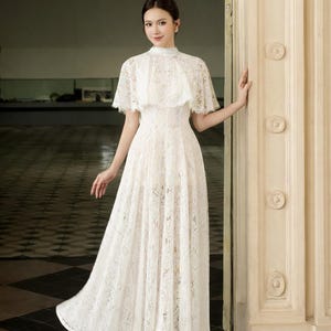 May include: A white lace dress with a high neckline and short cape sleeves. The dress has a fitted bodice and a flowing skirt. The woman is wearing white heels. The background is a doorway with decorative molding.